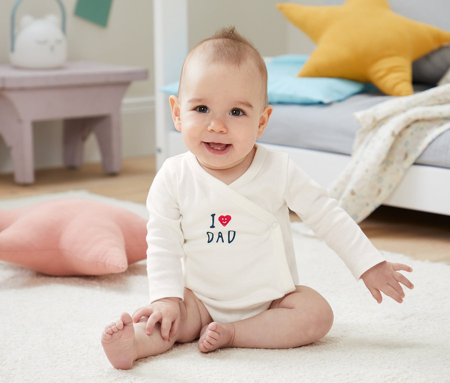 Usmievajúce sa dieťa sedí na bielom koberci a má na sebe biele body s dlhým rukávom z biobavlny s nápisom "I love Dad".