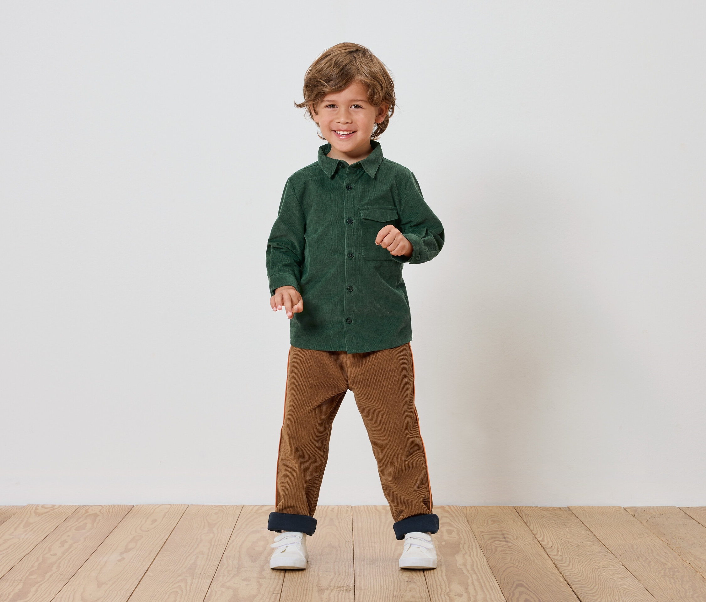 Chlapec stojí a usmieva sa, má na sebe zelenú košeľu, hnedé nohavice a biele topánky.