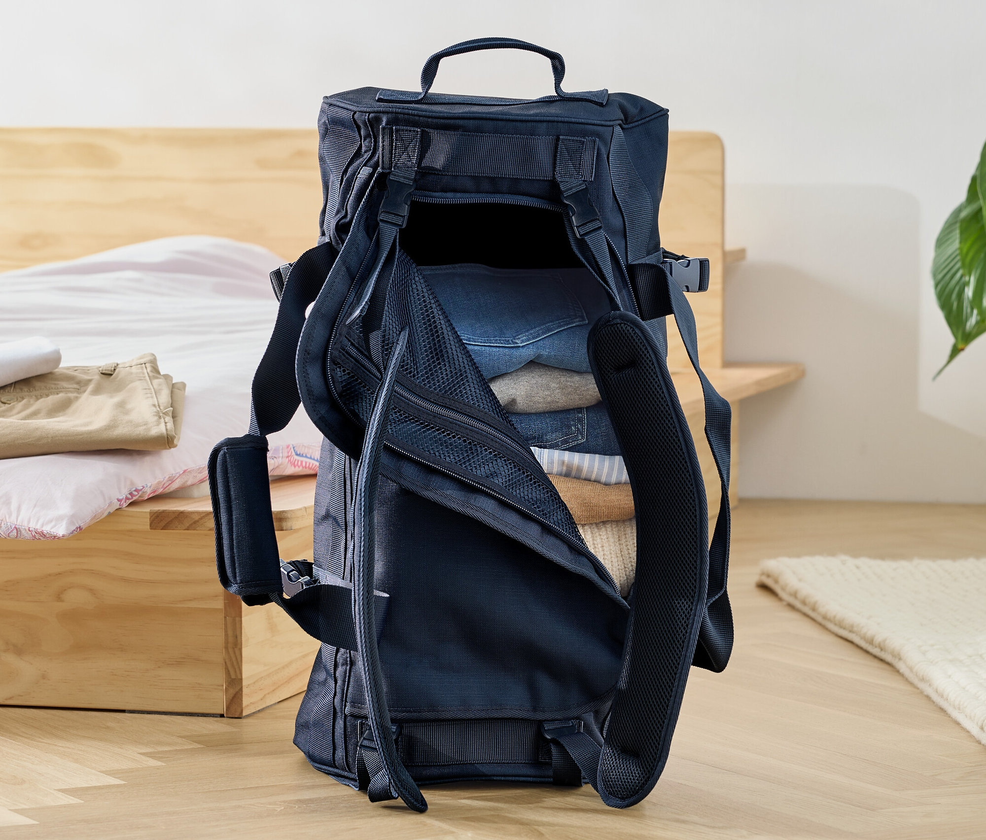 Otvorená cestovná taška a ruksak naplnená oblečením stojí na podlahe pri posteli.