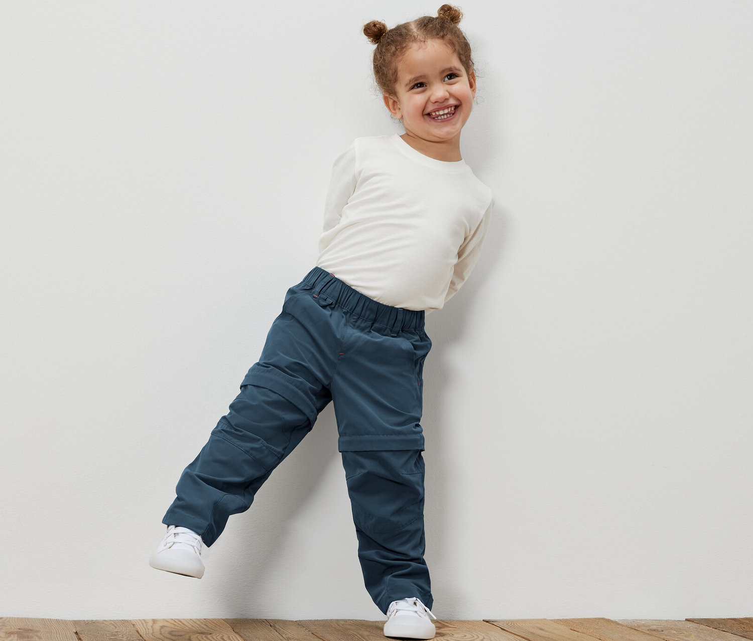 Usmievajúce sa dievčatko opreté o bielu stenu má na sebe funkčné nohavice pre malé deti a bielu košeľu.