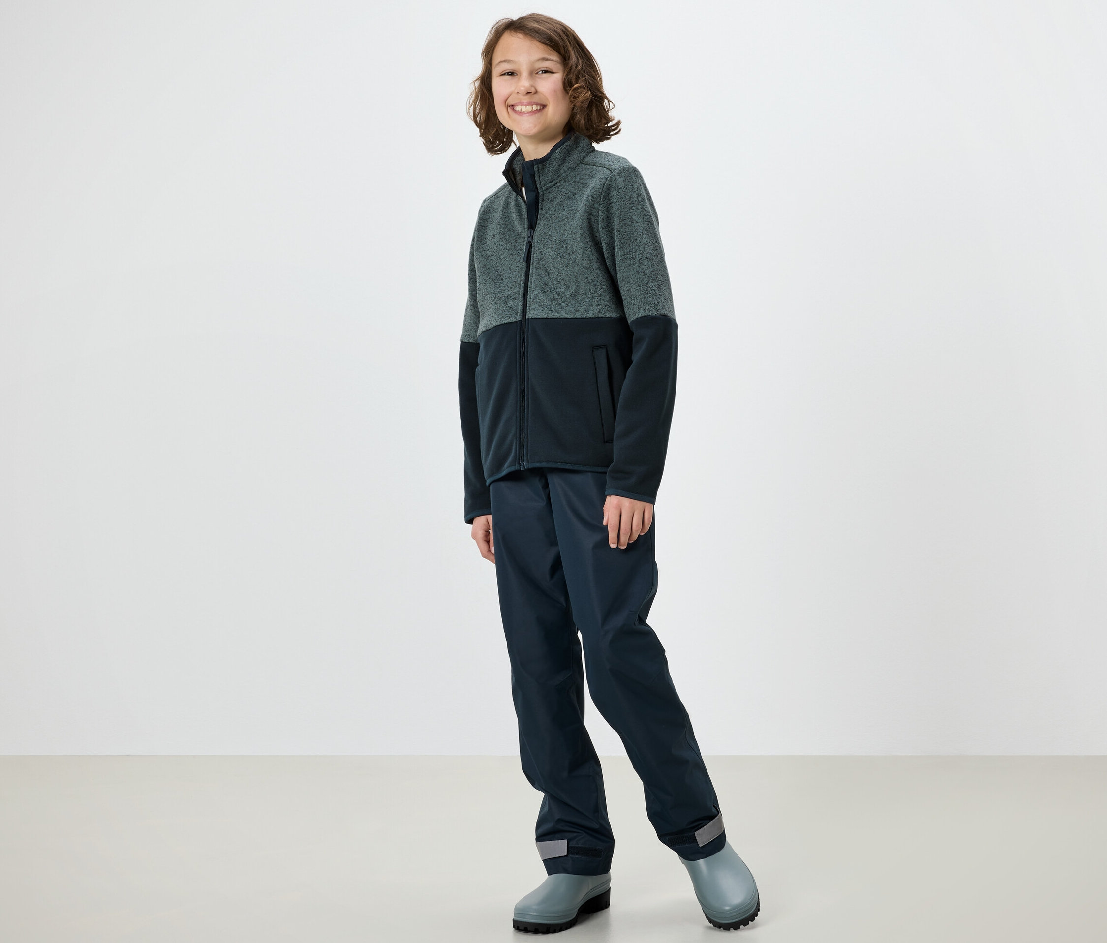 Dieťa pózuje v hybridnej pletenej fleecovej bunde, nohaviciach a gumákoch.