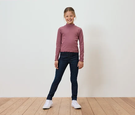 Usmievavé dievča v ružovom tričku s dlhým rukávom, tmavomodrých džeginách a bielych teniskách stojí na drevenej podlahe pred bielou stenou.
