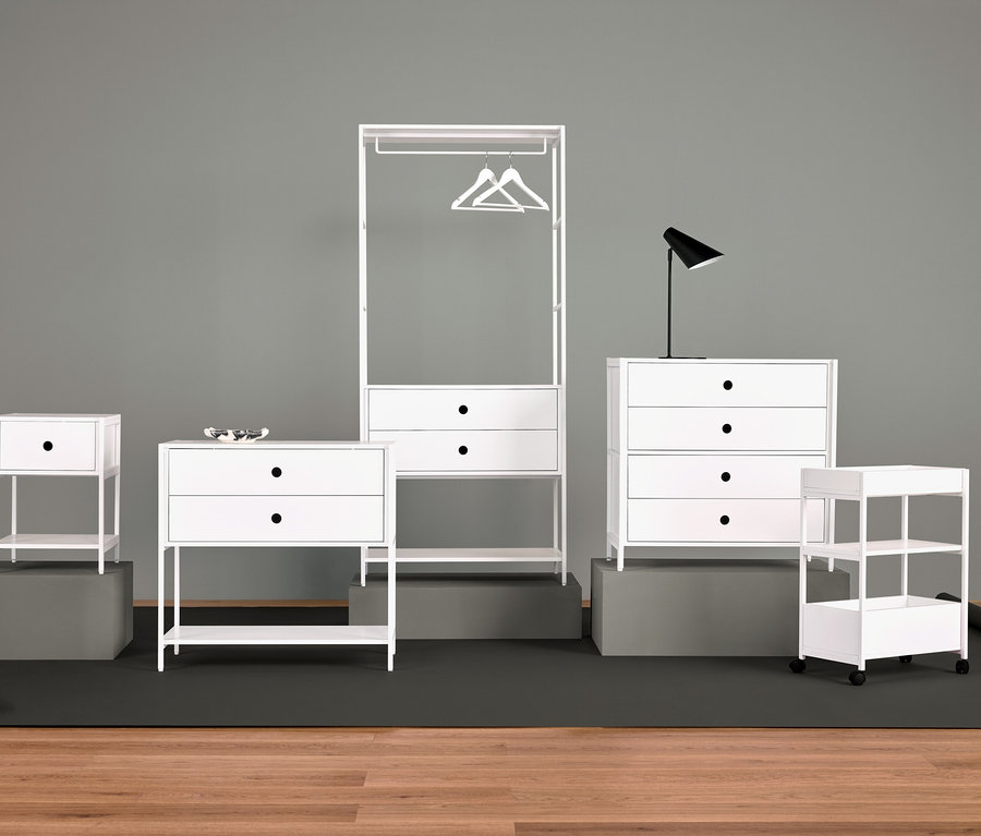 Usporiadanie bieleho nábytku s nočným stolíkom, komodou so zásuvkami, vešiakom na šaty, lampou a policami.