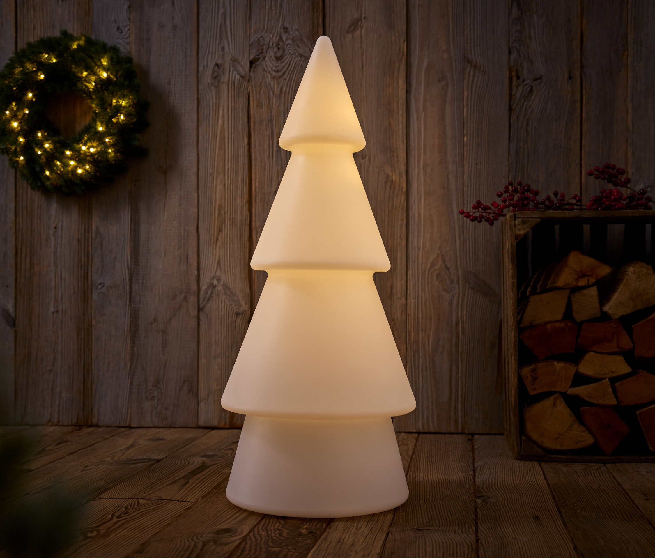 Zvislý záber osvetleného dekoratívneho vianočného stromčeka, vedľa ktorého je drevená debna s drevom a na stene visí veniec.