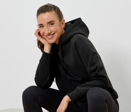 Usmievajúca sa žena v čiernej mikine s kapucňou a čiernych legínach sedí a opiera si tvár o ruku.