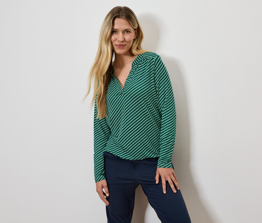 Žena s blond vlasmi pózuje v zelenej blúzke s prúžkami a tmavomodrých nohaviciach.