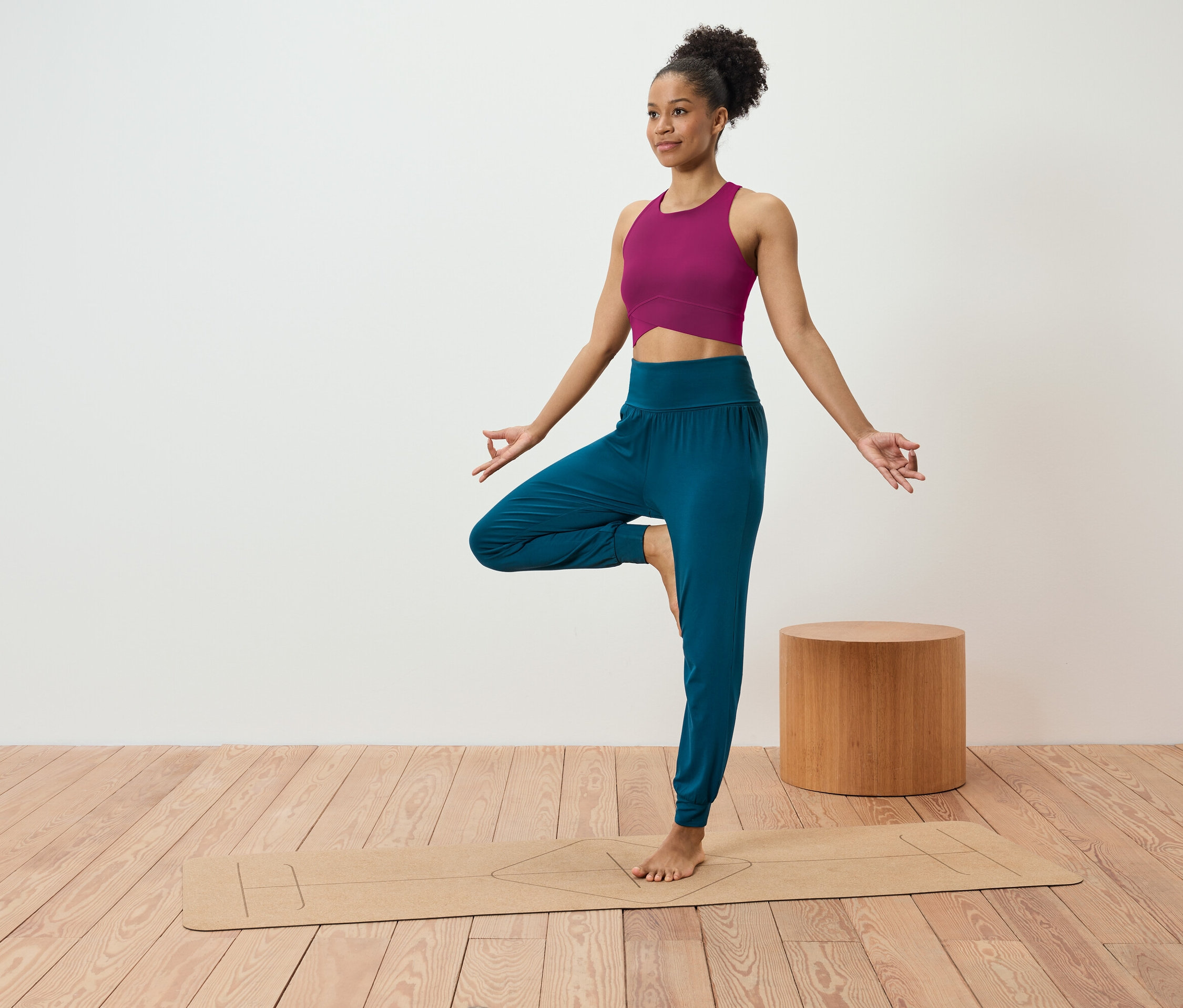 Žena v ružovom tope a modrých nohaviciach stojí v pozícii stromu na jogovej podložke.