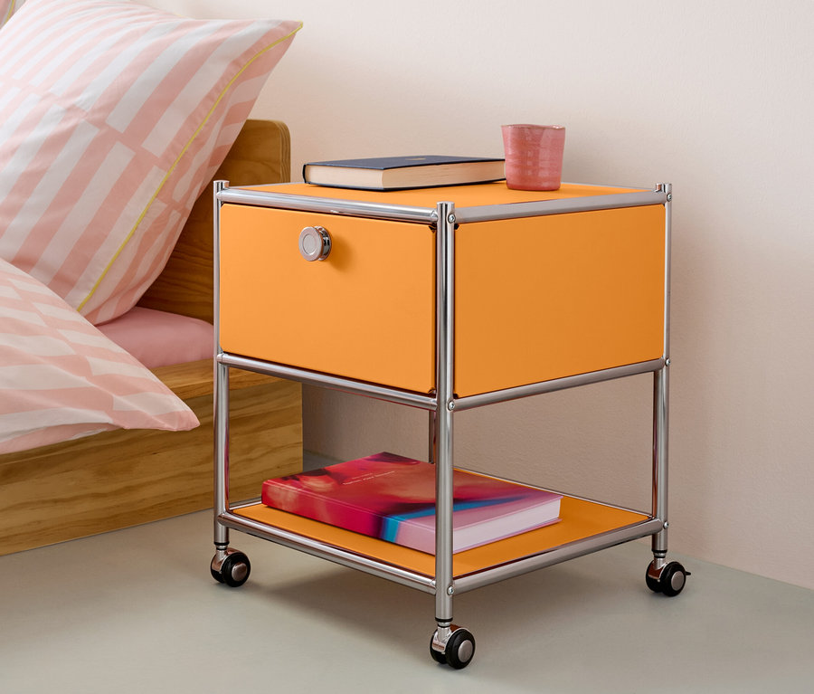 Oranžový kovový nočný stolík so zásuvkou »CN3« stojí vedľa postele.