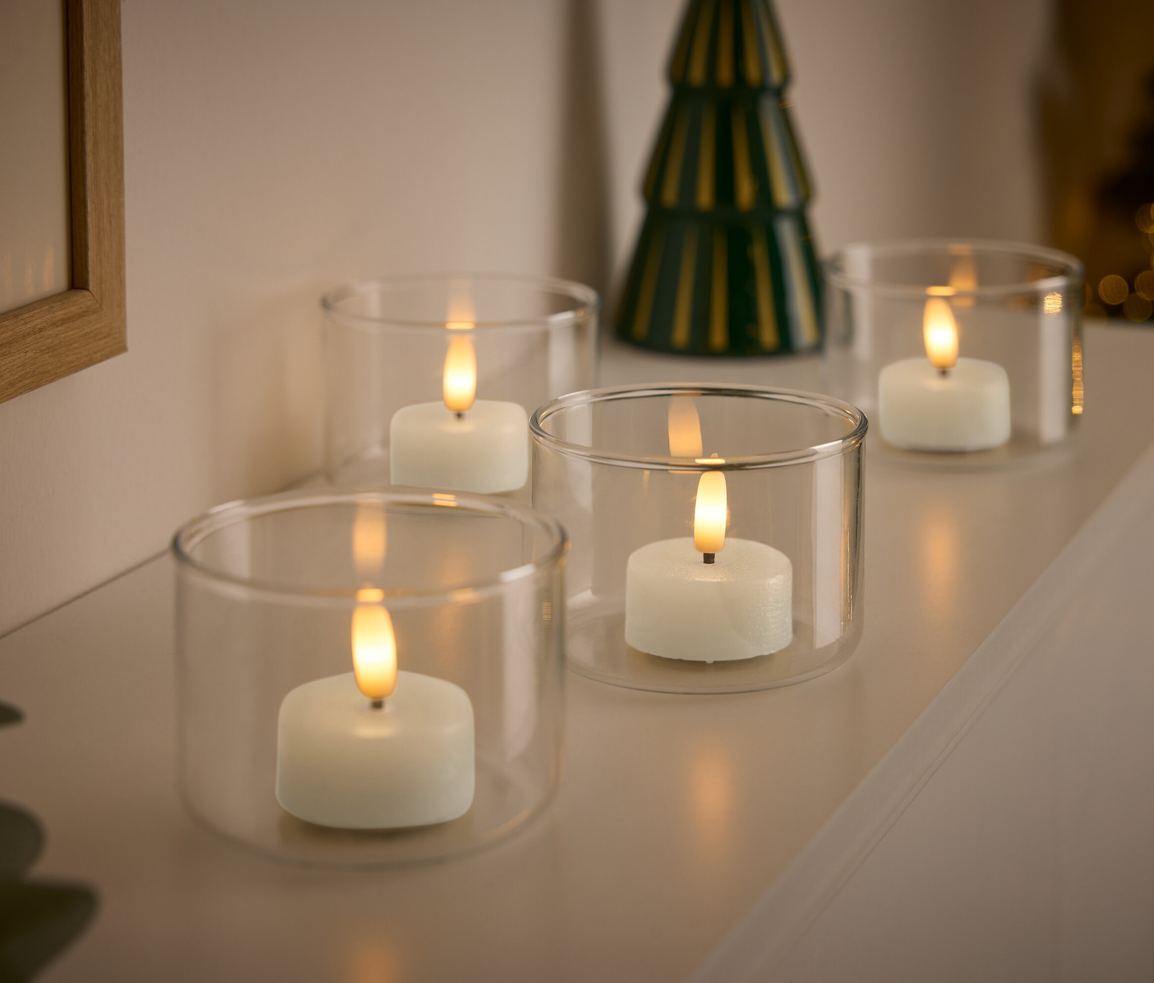 Štyri biele sviečky v priehľadných sklenených svietnikoch s malým zeleným vianočným stromčekom v pozadí.