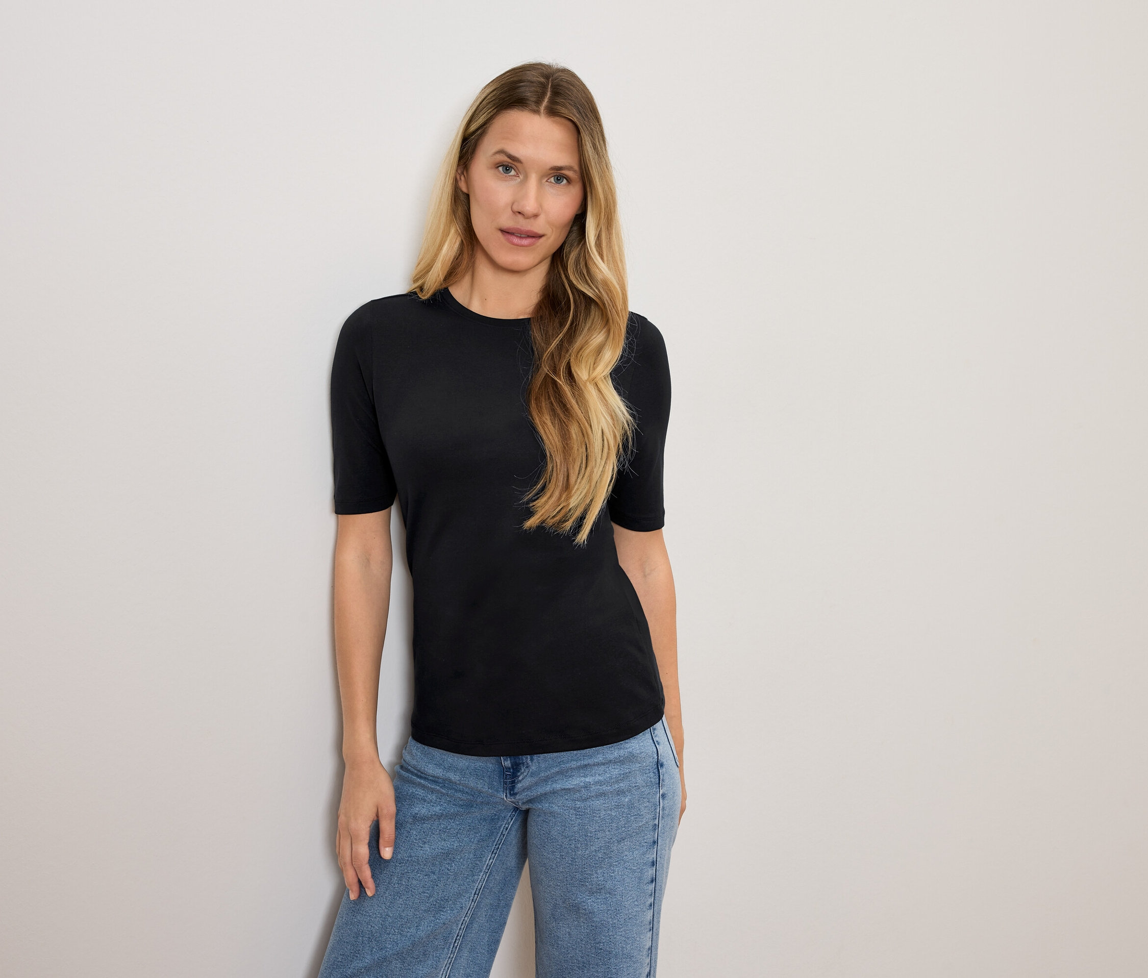 Žena s blond vlasmi pózuje v čiernom tričku a modrých džínsoch.