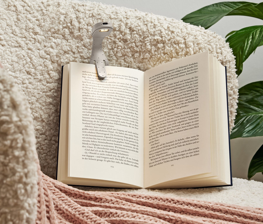 Sivá lampička na čítanie je pripevnená k otvorenej knihe ležiacej na mäkkej krémovej deke s ružovými akcentmi.