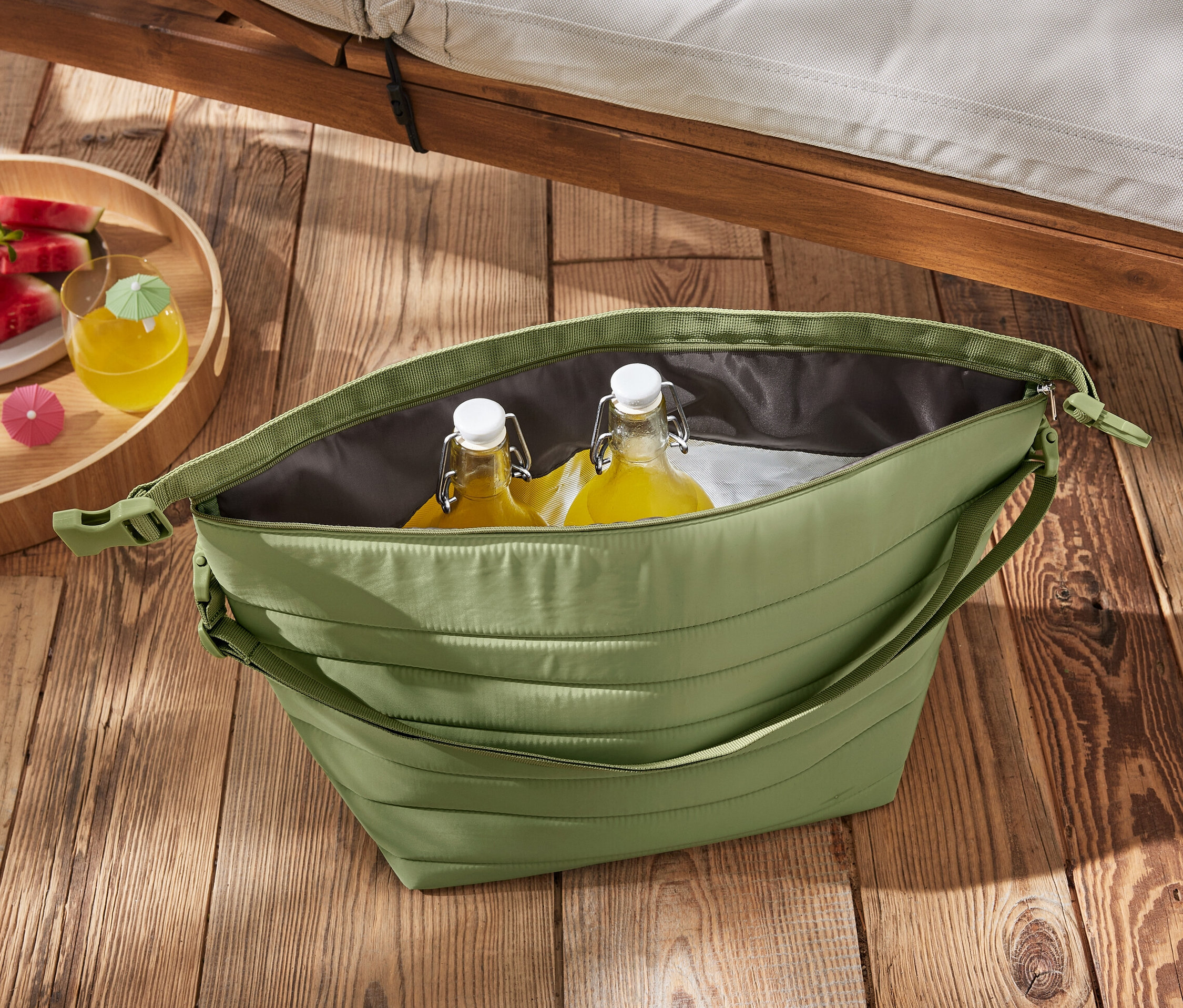 Zelená chladiaca taška s fľašami a melónom na drevenom stole.