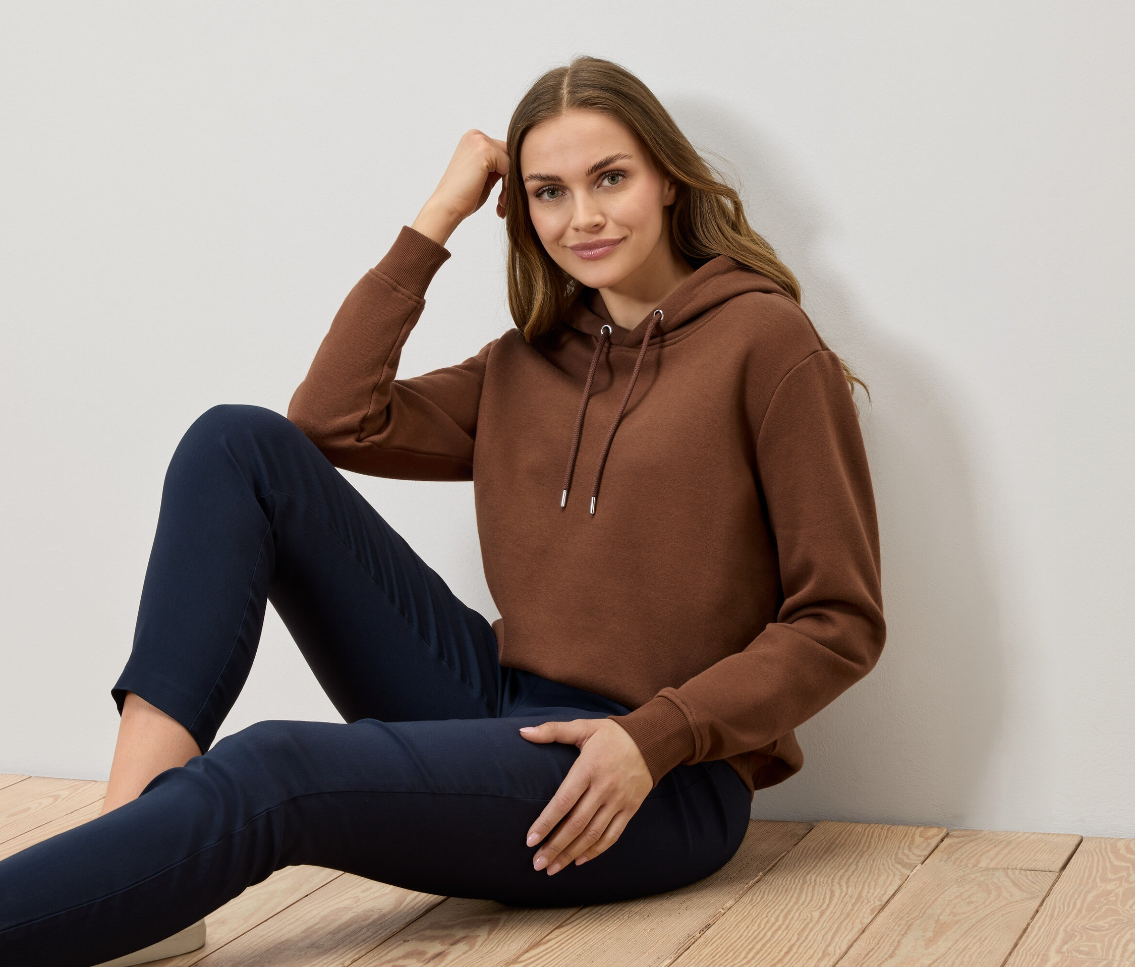 Žena sedí na drevenej podlahe a má na sebe hnedú mikinu a tmavomodré nohavice.