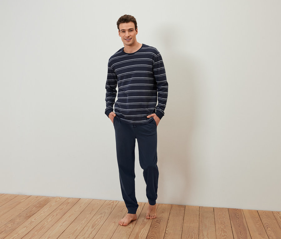 Muž stojí bosý a má ruky vo vreckách tmavomodrého pruhovaného pyžama.