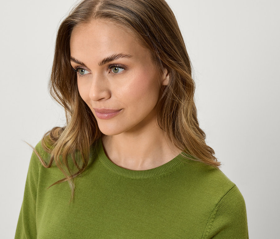 Portrét ženy s hnedými vlnitými vlasmi a zeleným svetrom, pozerá sa nabok.