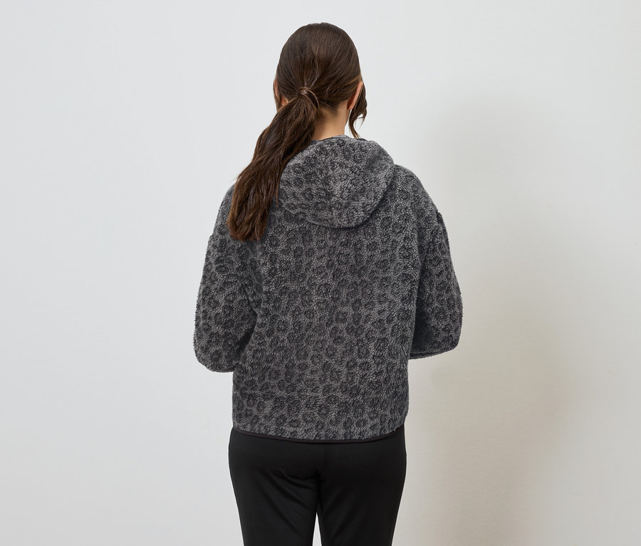 Žena chrbtom k fotoaparátu má na sebe sivú mikinu s kapucňou so zvieracím vzorom a čierne nohavice.