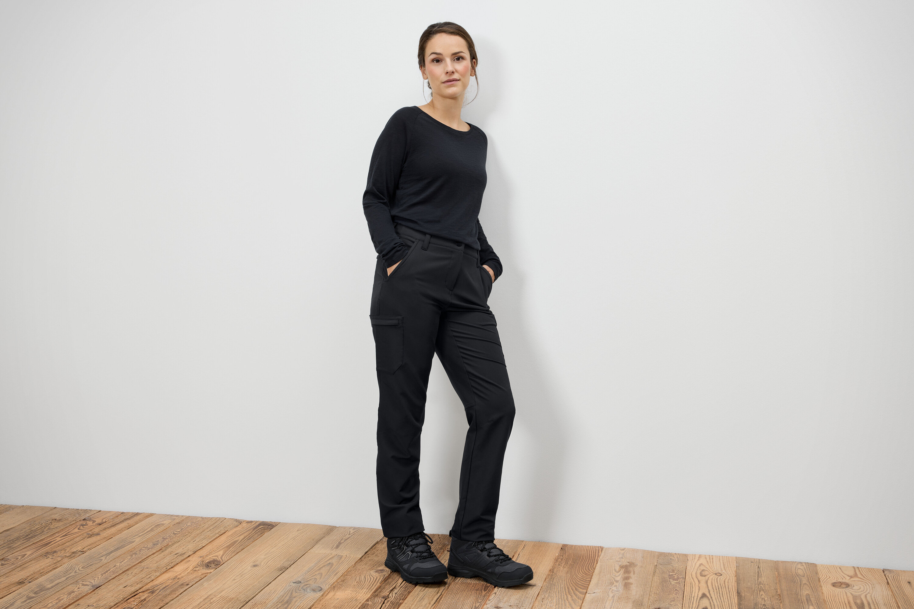 Žena stojí s rukami vo vreckách v čiernych nohaviciach a čiernych topánkach.