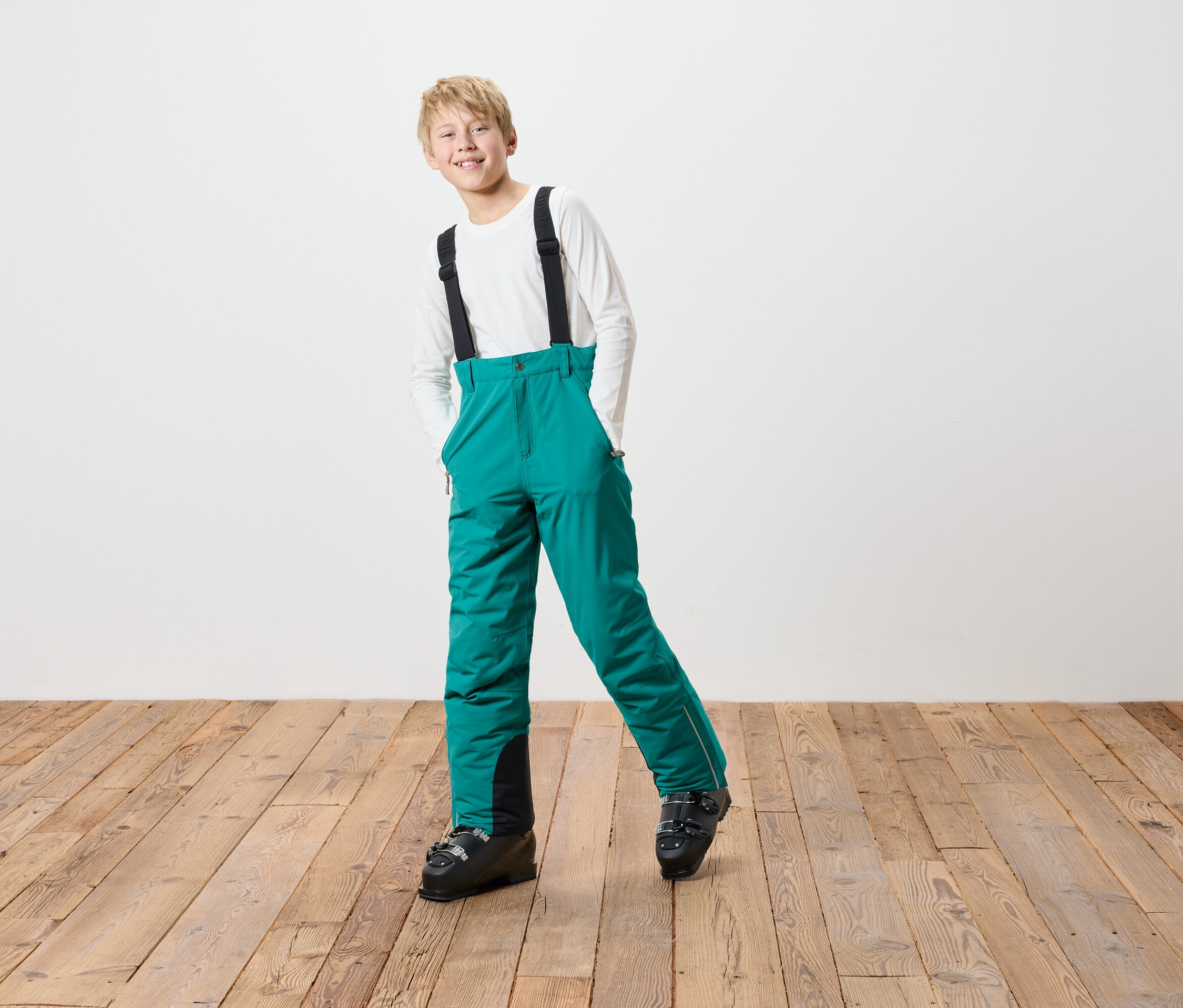 Chlapec v zelených lyžiarskych nohaviciach s čiernymi trakami a lyžiarskych topánkach stojí na drevenej podlahe pred bielou stenou.