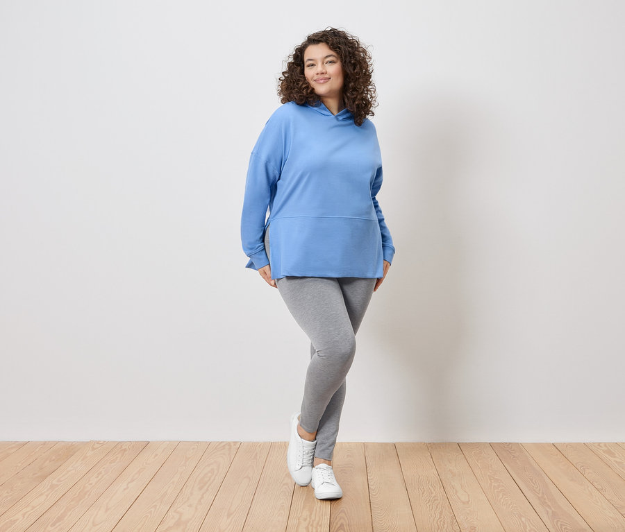 Žena s kučeravými vlasmi stojí pred bielou stenou a má na sebe modrú mikinu s kapucňou, sivé legíny a biele tenisky.