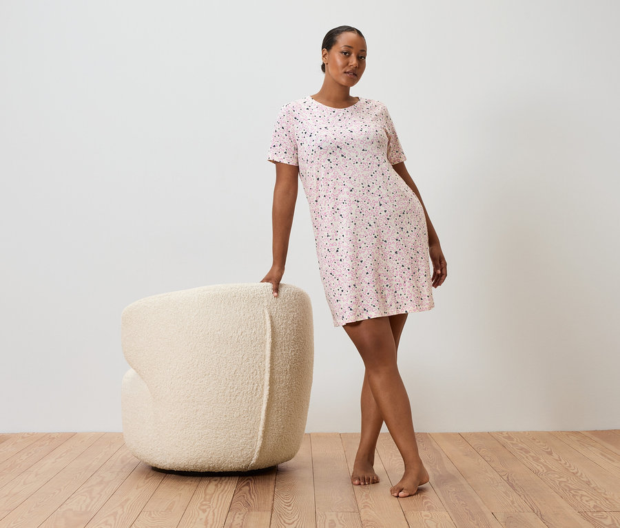 Žena sa opiera o krémové kreslo, má na sebe ružové bodkované šaty s krátkym rukávom.