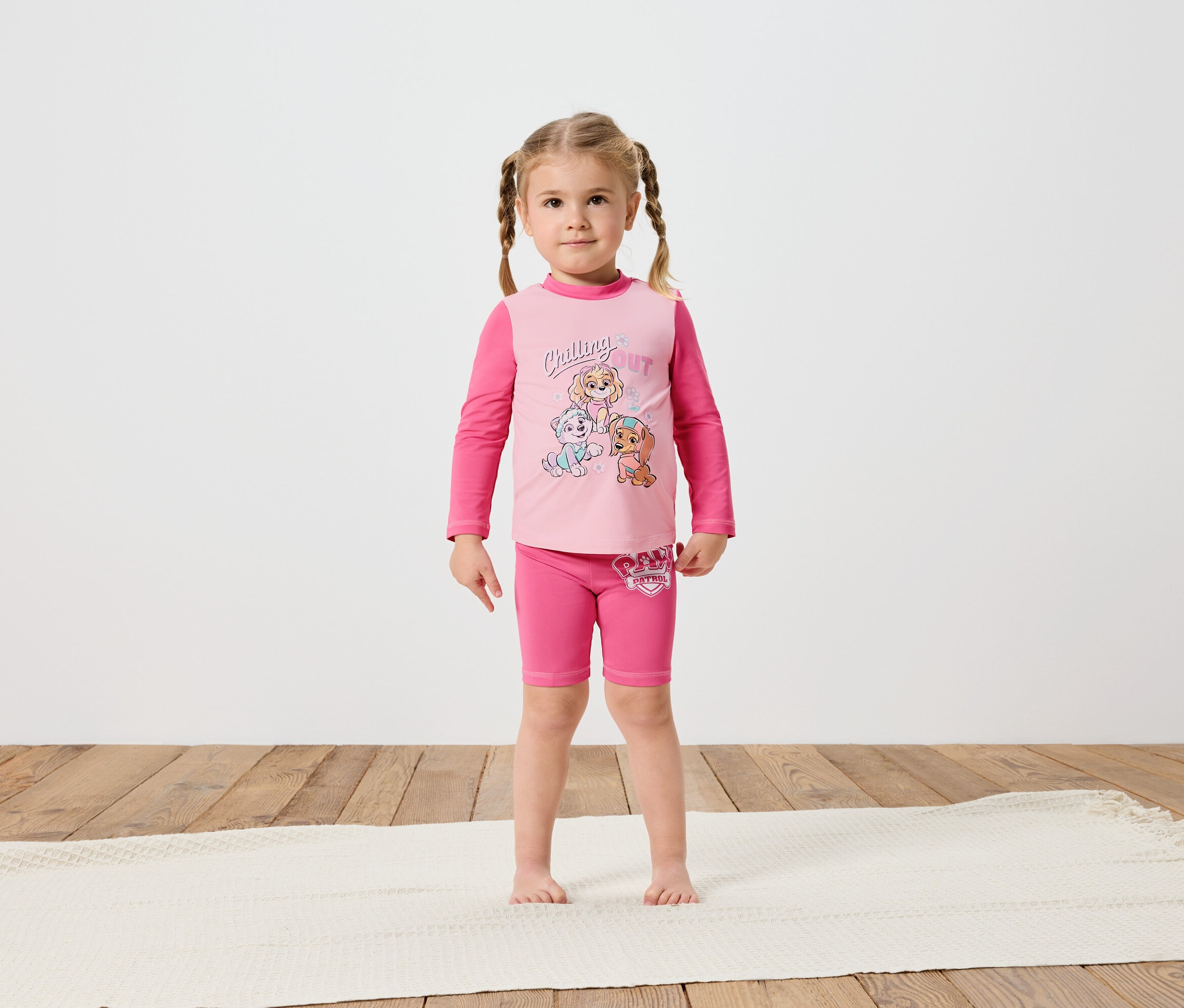 Malé dievčatko so zapletenými vlasmi stojí bosé na krémovom koberci a má na sebe ružové tričko s dlhým rukávom a šortky Paw Patrol.