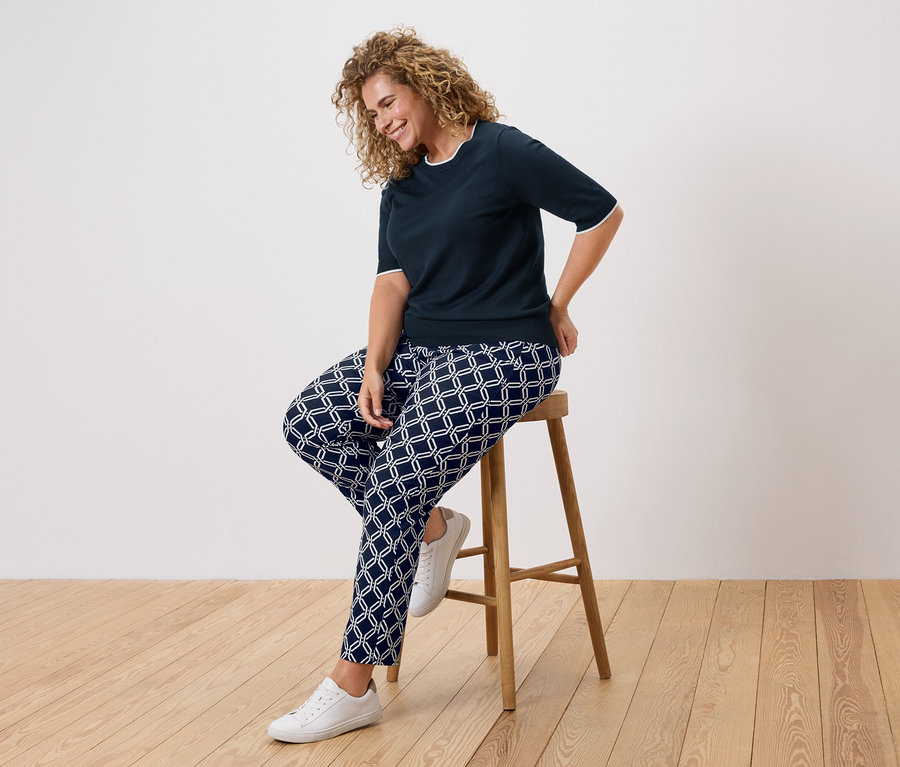 Žena s kučeravými vlasmi sedí na stoličke, oblečená v tmavomodrej blúzke a vzorovaných nohaviciach.