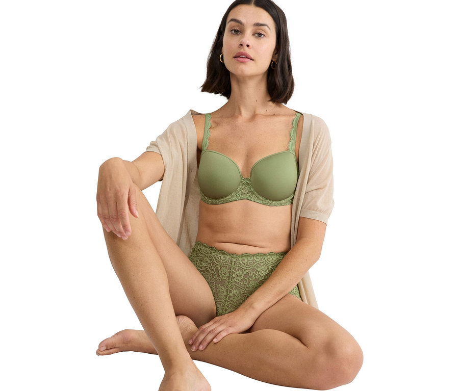 Žena sedí so skríženými nohami a má na sebe zelenú podprsenku a zelené čipkované nohavičky, spolu s béžovým kardigánom.