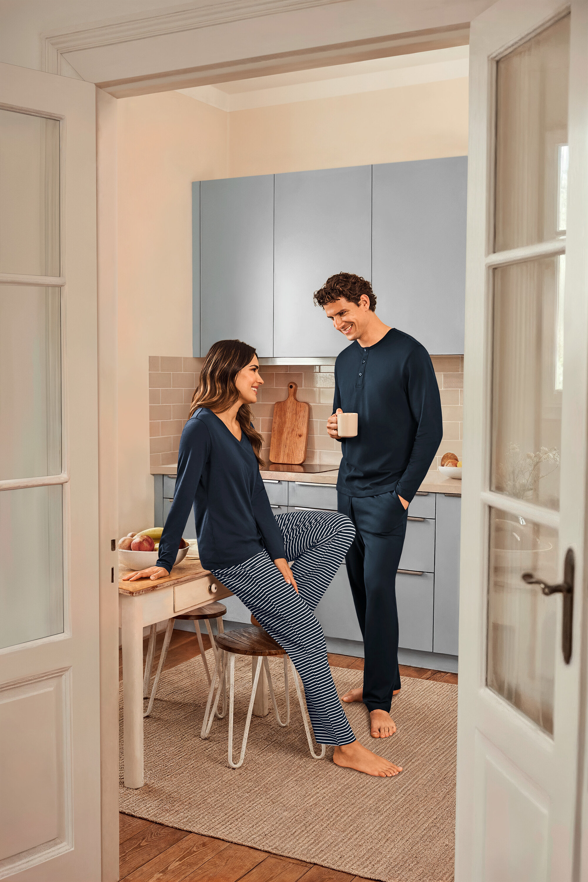 V kuchyni stojí žena sediaca na stole v tmavomodrom prúžkovanom pyžame a tmavomodrom vrchnom diele a muž, ktorý drží šálku a má na sebe tmavomodré pyžamo.