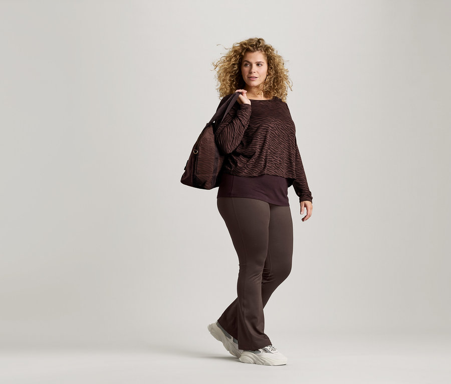 Žena s kučeravými vlasmi má na sebe hnedú blúzku so zvieracou potlačou, hnedé legíny a tenisky. Cez rameno má prehodenú hnedú tašku.