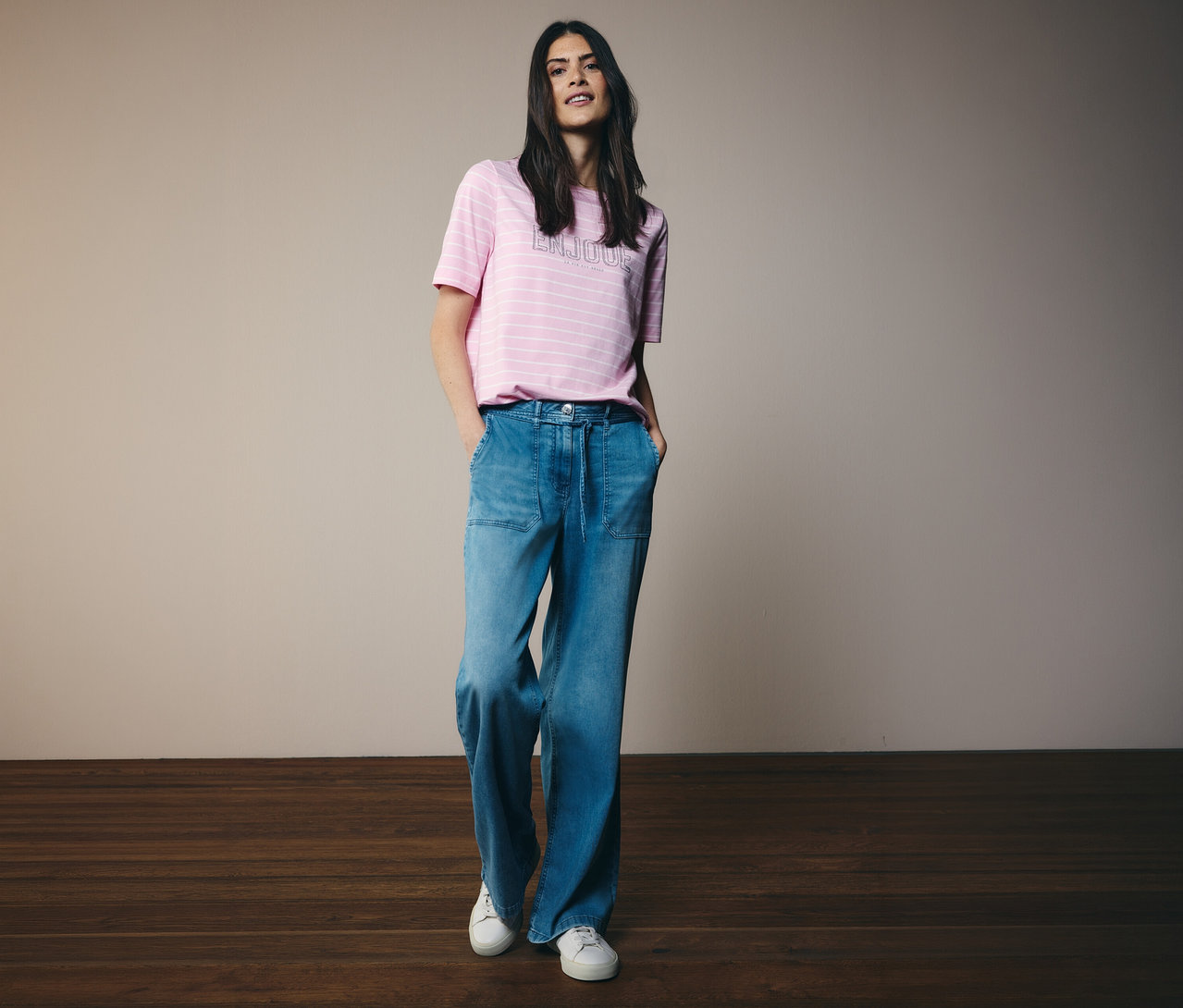 Žena v tričku CECIL s ružovými pruhmi a širokých džínsoch a bielych teniskách pózuje s rukami vo vreckách.