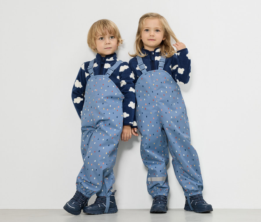 Dve deti stoja vedľa seba, chlapec a dievča, obaja majú na sebe modré flísové bundy s obláčikovým vzorom a modré nepremokavé traky s farebnými bodkami.