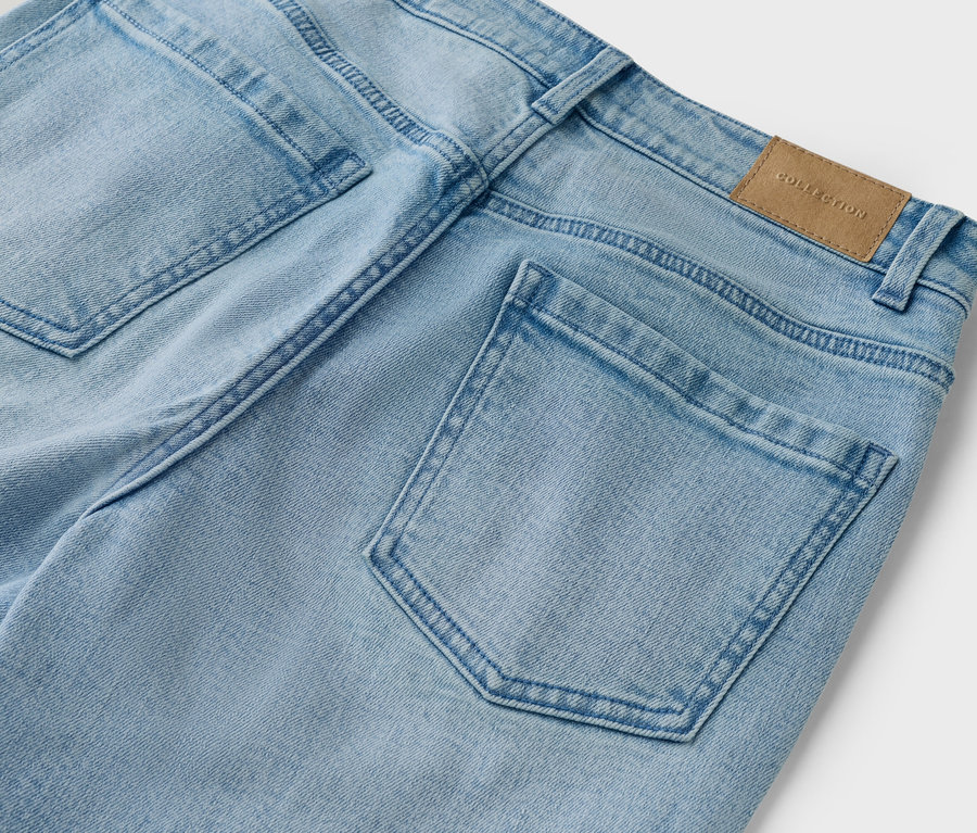 Detailný záber na zadnú časť svetlomodrých džínsov s vreckami a hnedým štítkom 'Collection'.