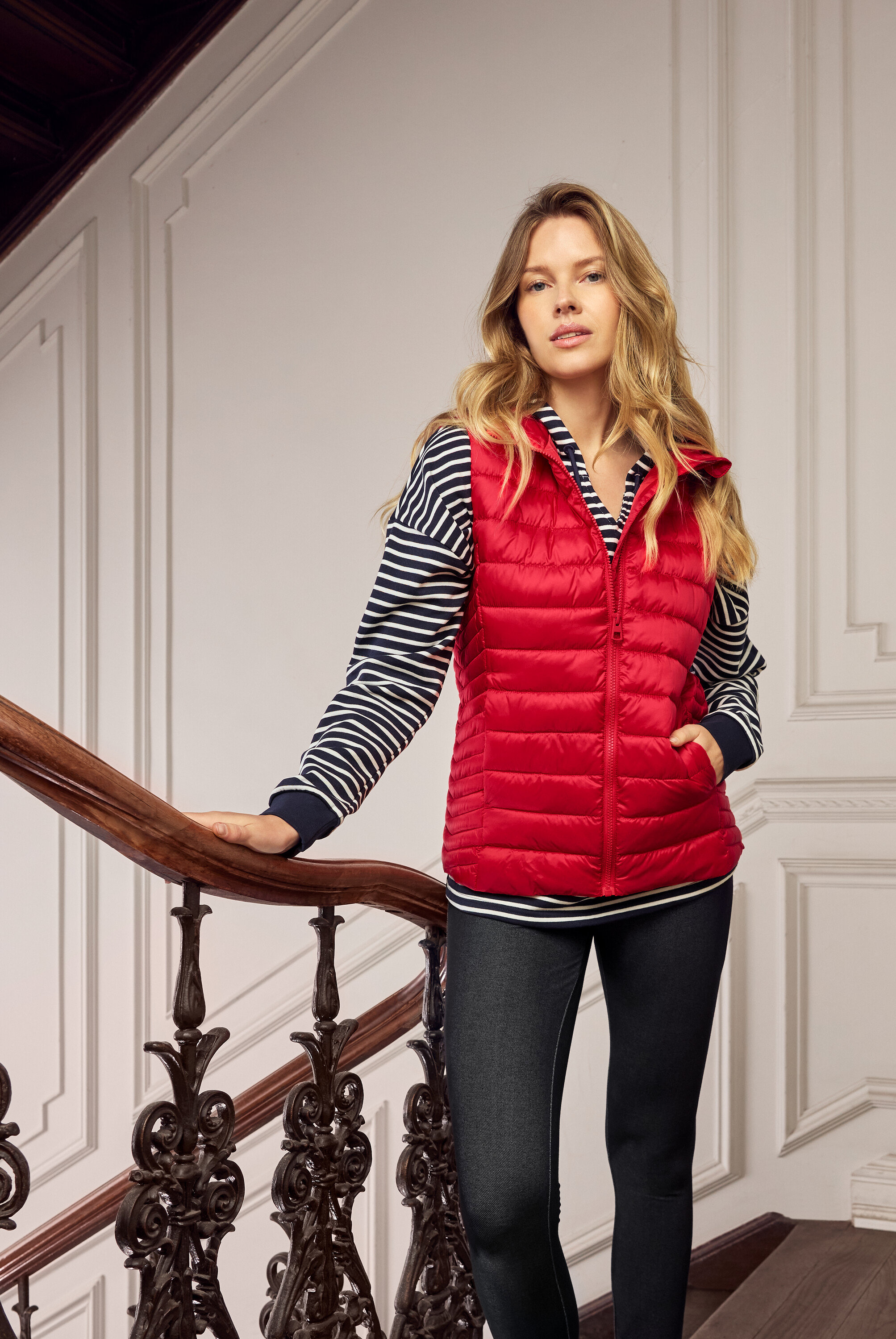 Žena s blond vlasmi v červenej prešívanej veste, modro-bielom pruhovanom tope a tmavých džínsoch na schodisku.