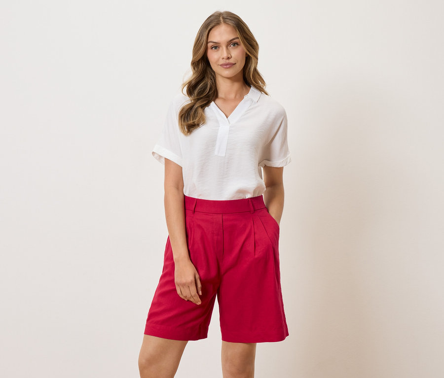 Žena v bielej blúzke a červených šortkách stojí na svetlom pozadí.