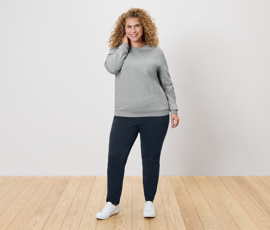 Žena s kučeravými vlasmi v sivom svetri a tmavomodrých nohaviciach.