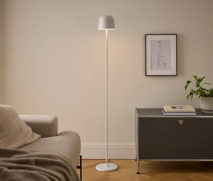 Moderná obývačka s béžovou pohovkou, bielou stojacou lampou a tmavosivou skrinkou. Osvetlená lampa stojí medzi pohovkou a skrinkou, na ktorej sú knihy a črepníková rastlina pod zarámovaným obrazom.