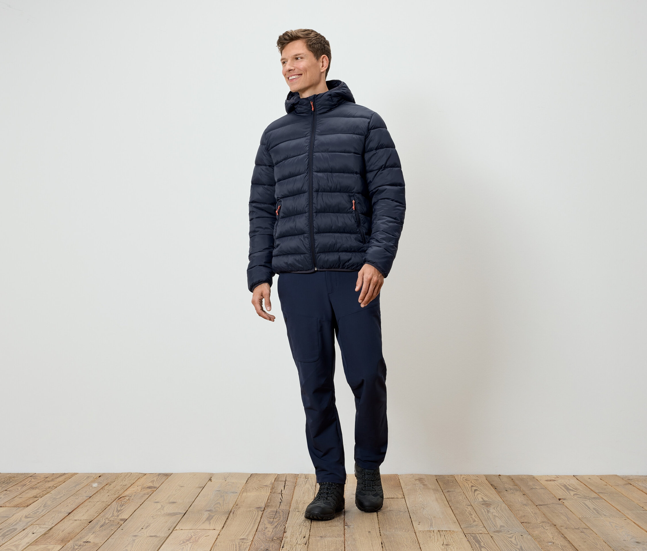 Muž stojí a má na sebe tmavomodrú prešívanú bundu, tmavomodré nohavice a čierne topánky.