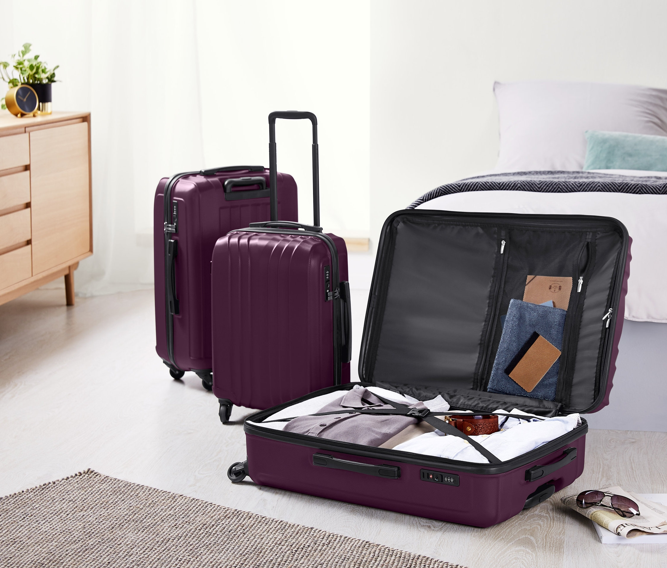 Tri fialové pevné škrupinové kufre (malý, stredný a veľký) stoja v izbe vedľa postele a komody. Jeden kufor je otvorený a plný oblečenia.