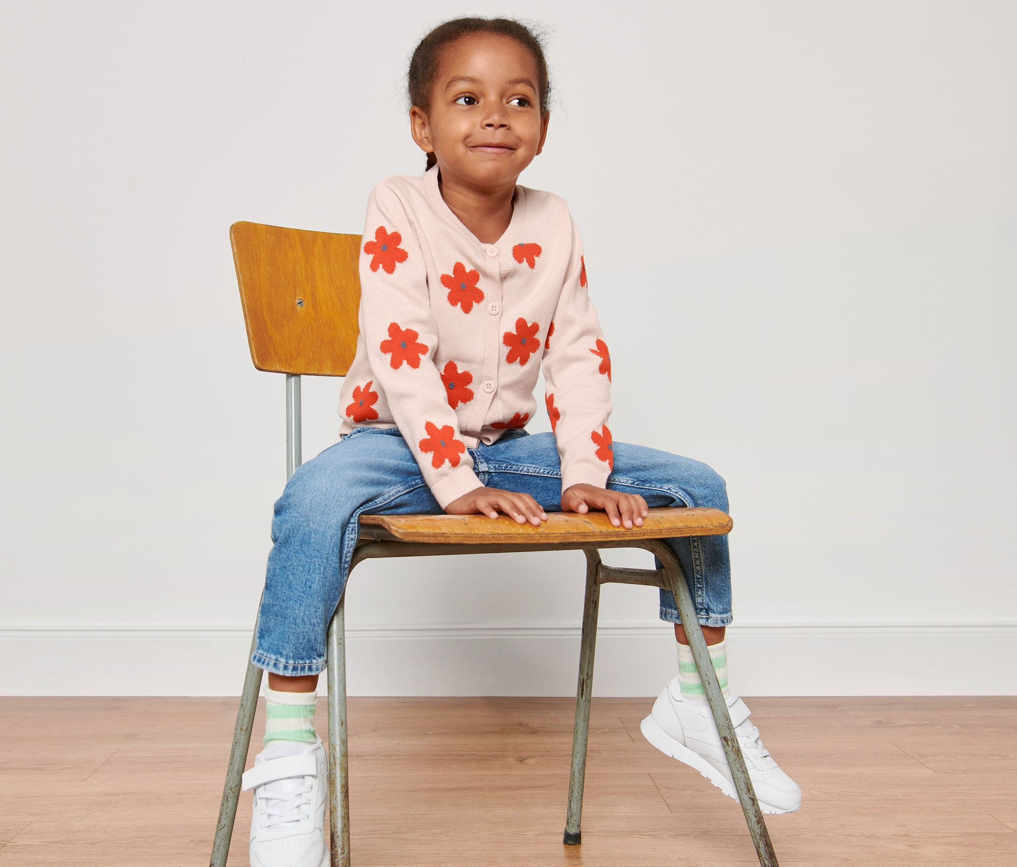 Dievča sedí na stoličke a má na sebe detský pletený sveter s kvetinovým vzorom.