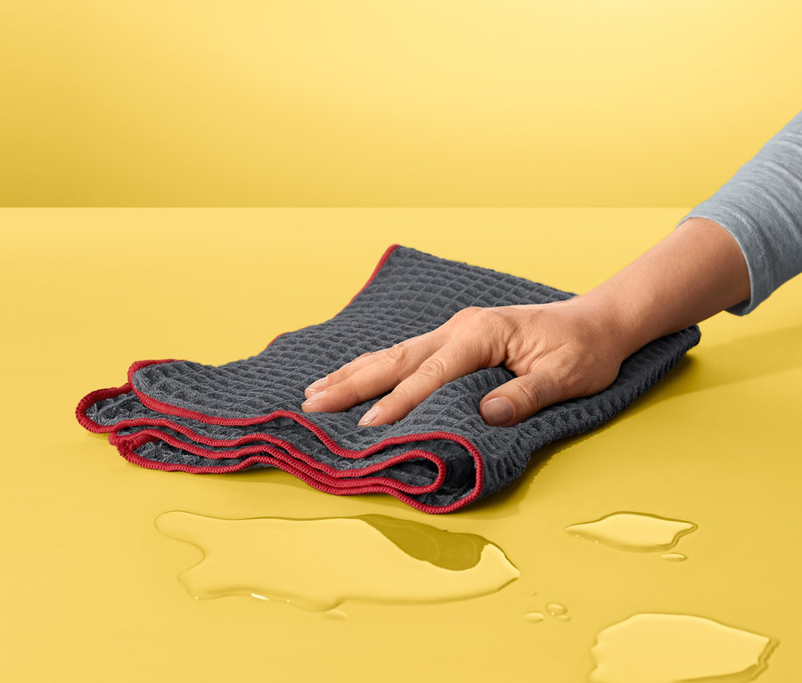 Ruka utiera rozliatu vodu pomocou Špeciálnej handričky na utieranie dosucha na žltom povrchu.