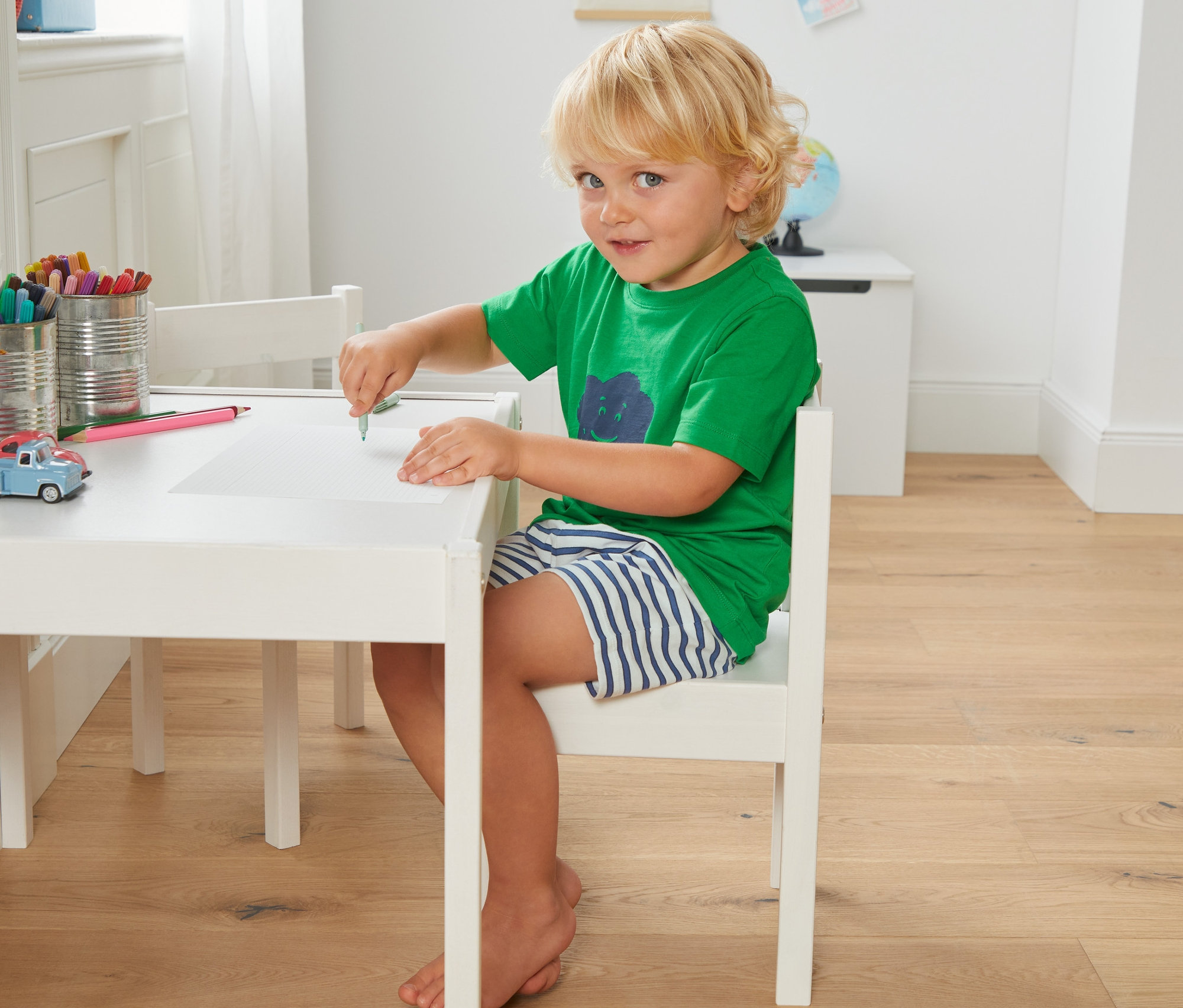 Chlapec sedí za stolom a kreslí na papier. Má na sebe zelené tričko a krátke pruhované pyžamo z biobavlny, 2 ks.