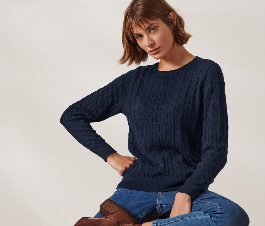 Žena sedí na stoličke a má na sebe tmavomodrý pletený pulóver s vrkočovým vzorom.