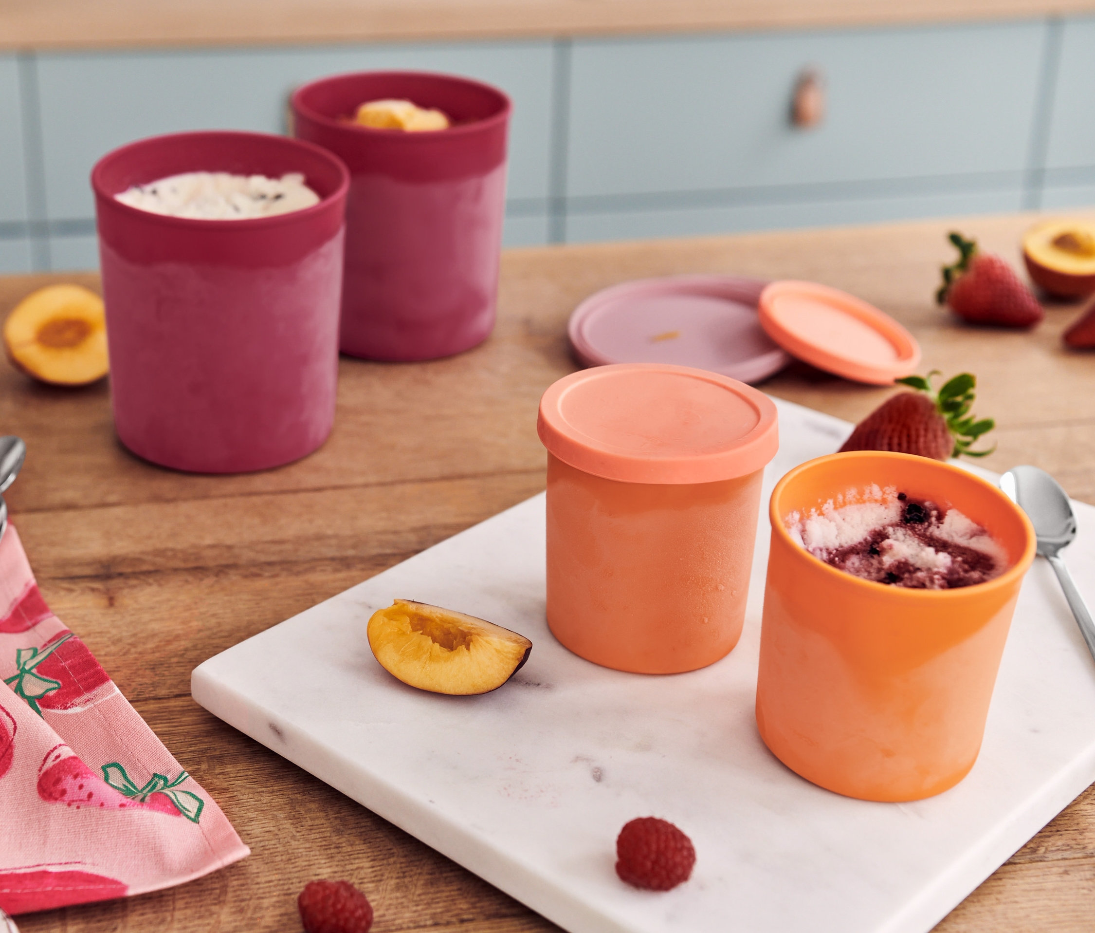 Dve červené a dve oranžové dózy do mrazničky s ovocím a lyžičkami na stole s utierkou.