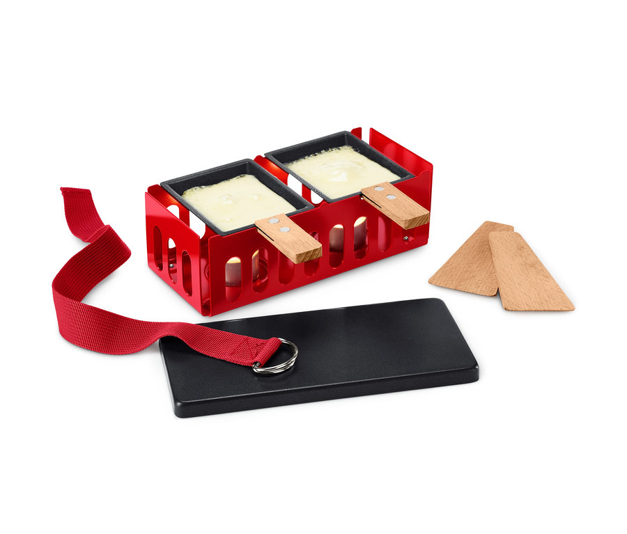 Súprava na Raclette pre dvoch s červeným rámom, dvoma panvicami s roztaveným syrom, čiernou doskou a drevenými špachtľami.