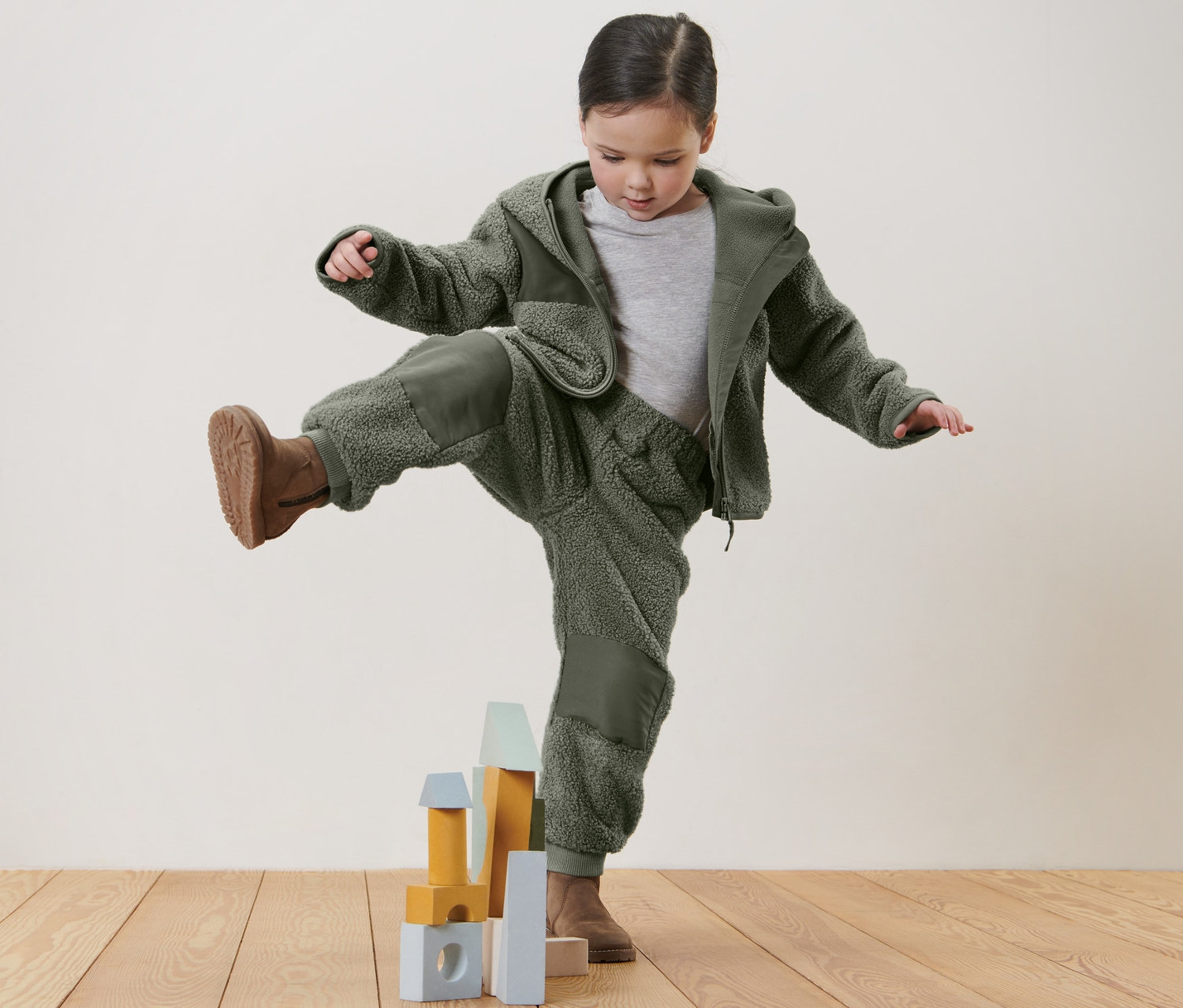 Dievča stojí na jednej nohe na drevenej podlahe a hrá sa s detskými mäkučkými nohavicami a kockami.