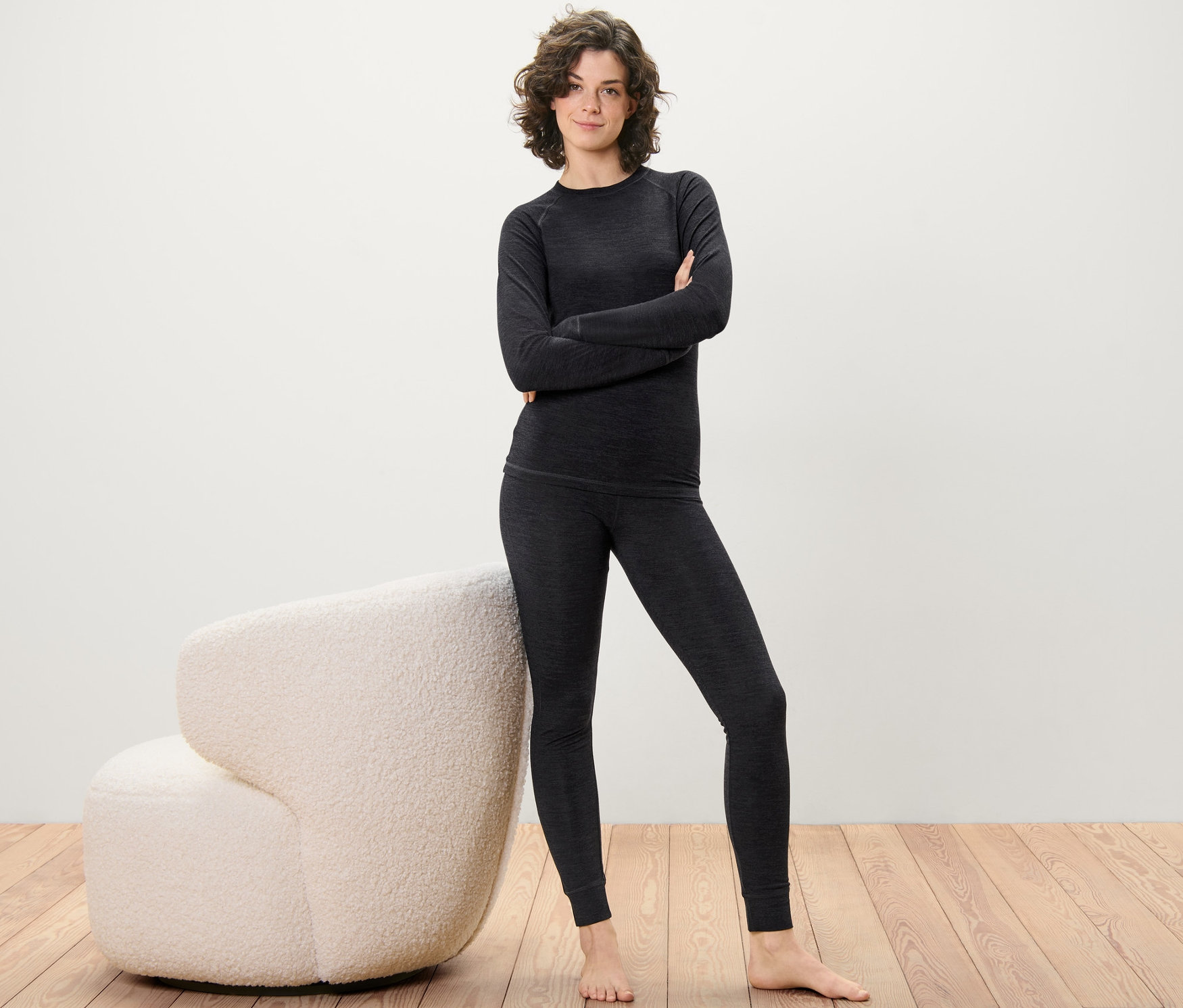 Žena pózuje v dlhých spodkoch s vlnou s certifikátom RWS a vrchnom diele na drevenej podlahe vedľa krémového kresla.