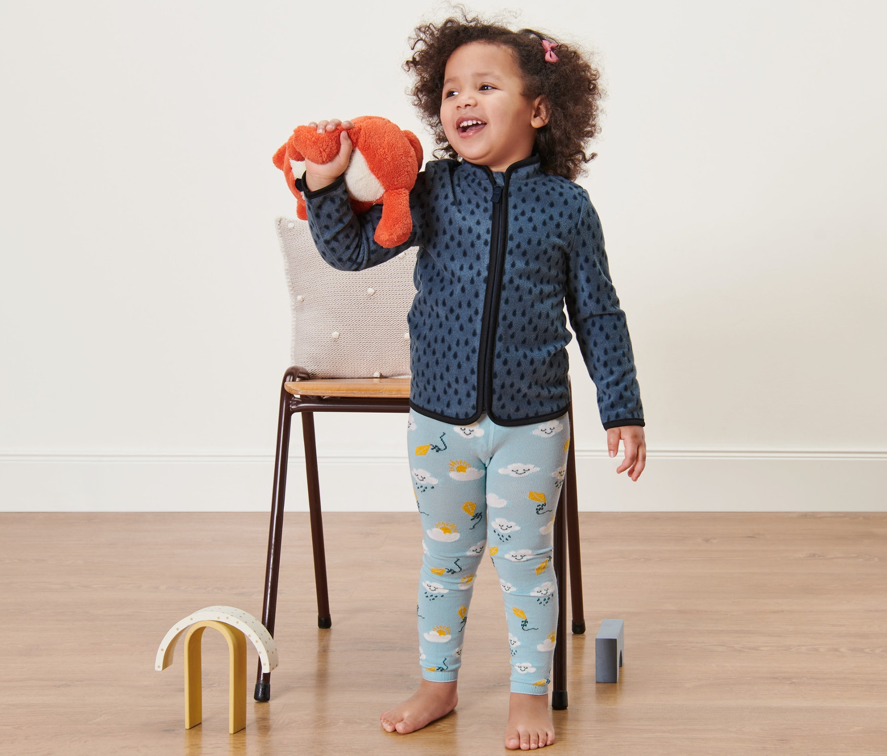 Usmievajúce sa dievčatko drží plyšovú líšku. Má na sebe modré legíny s obláčikmi a modrú mikinu.