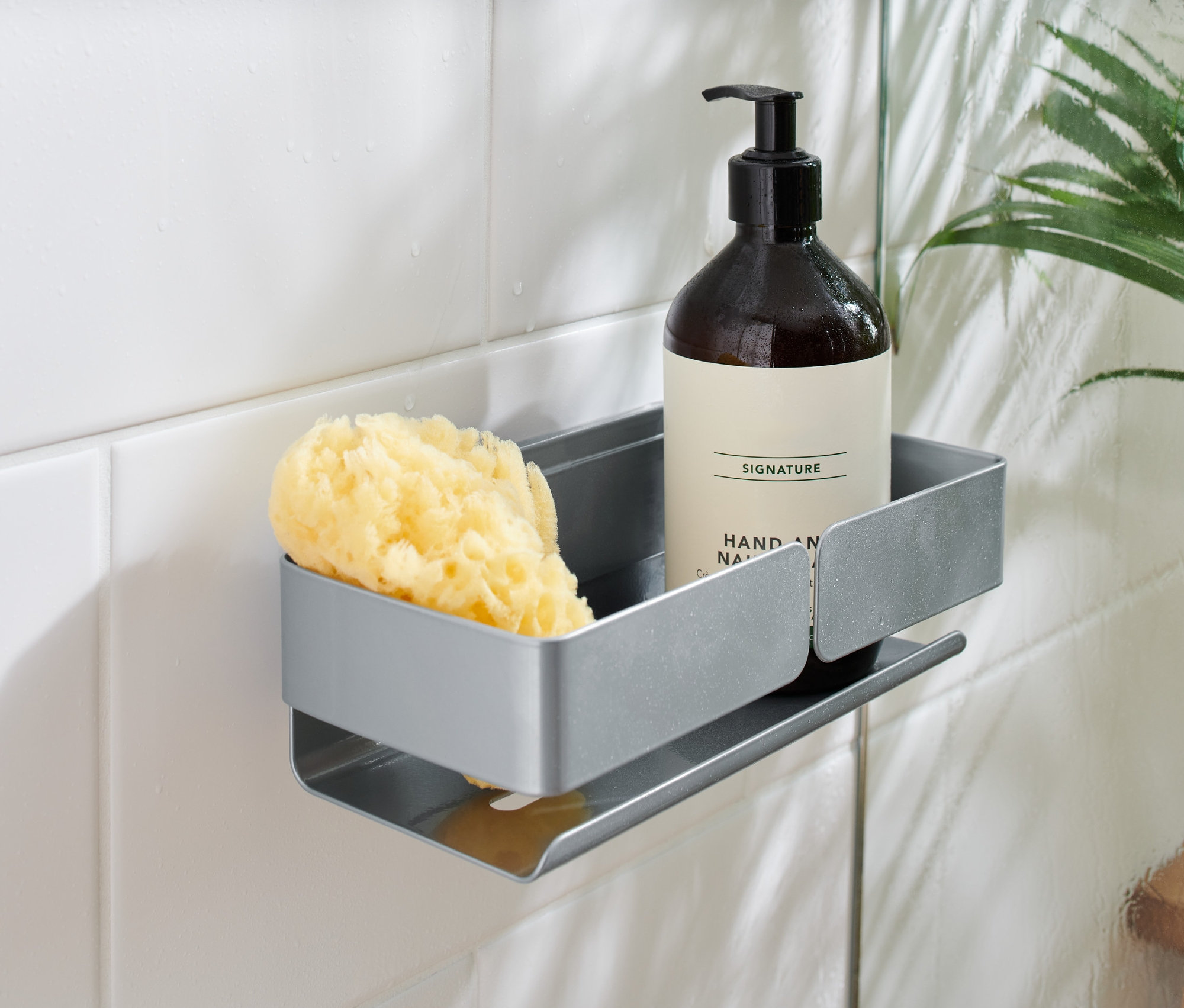 Sivá polička do sprchy pripevnená na bielych kachličkách drží hnedú fľaštičku s pumpičkou a žltú špongiu.