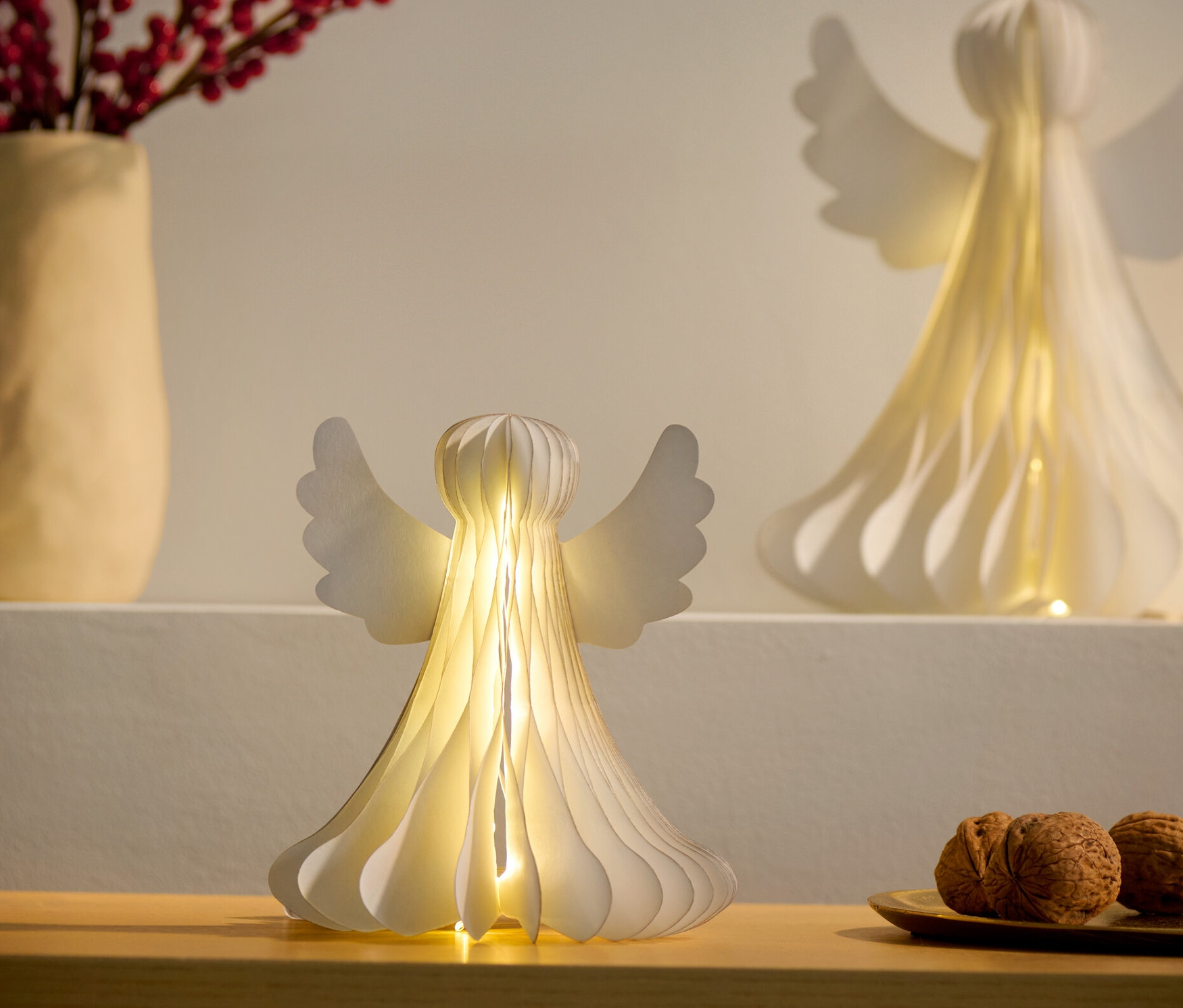 Dva malé papierové LED anjeli, jeden na skrinke a druhý na poličke, vedľa vázy s červenými bobuľami a taniera s orechmi.