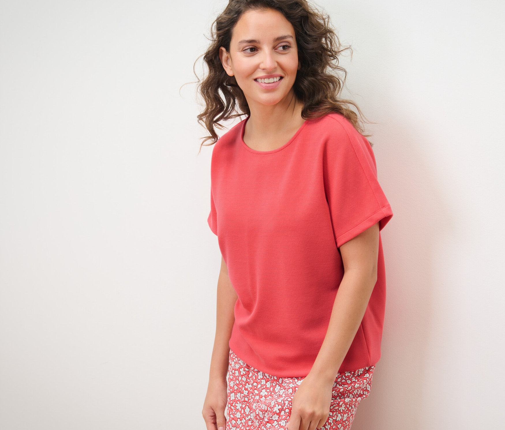 Žena pózuje na bielom pozadí a má na sebe koralové blúzkové tričko.